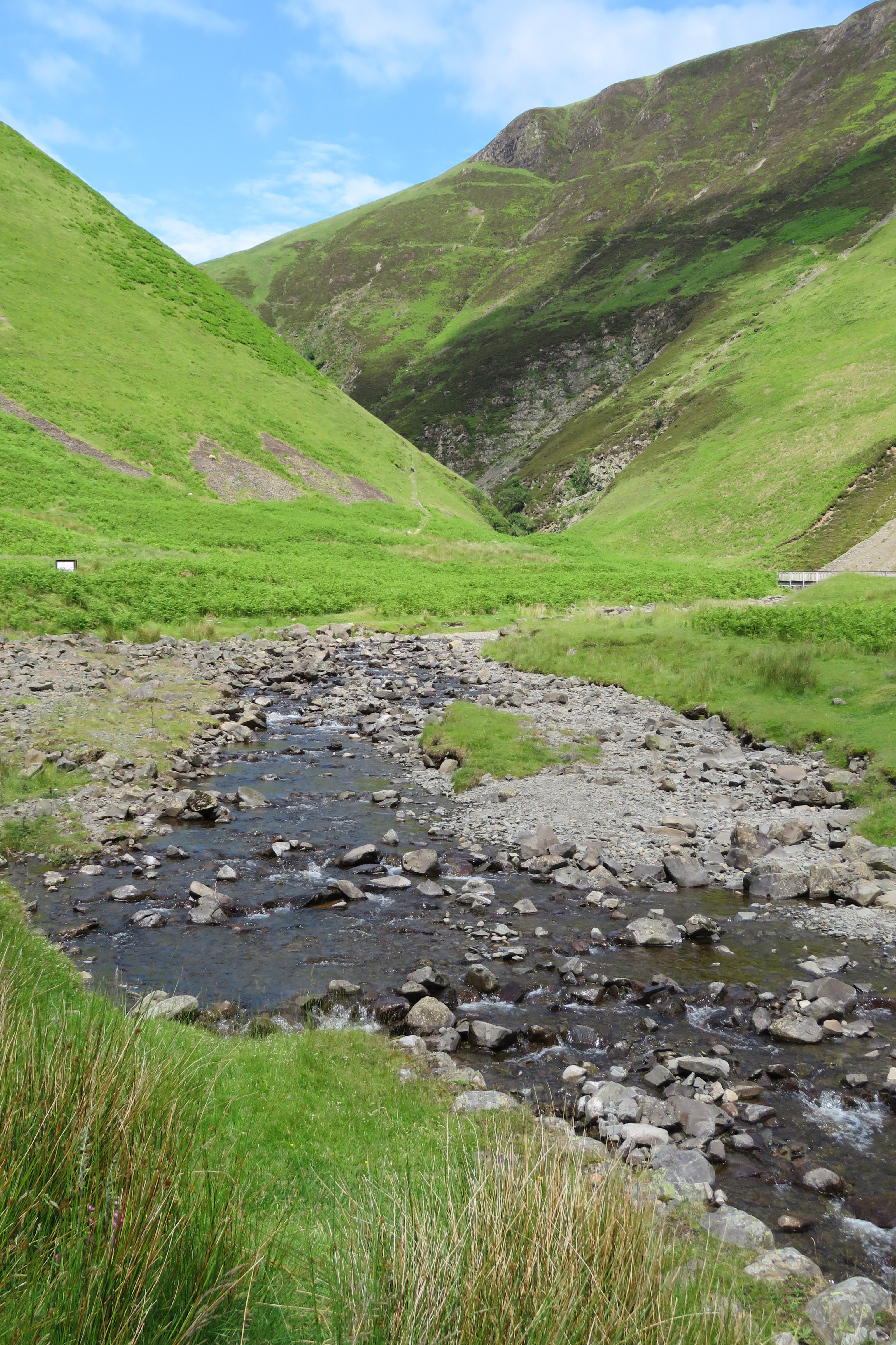 Rocky stream flowing through a green valley in the Scottish Highlands, with steep grassy hillsides rising on both sides under a partly cloudy blue sky.